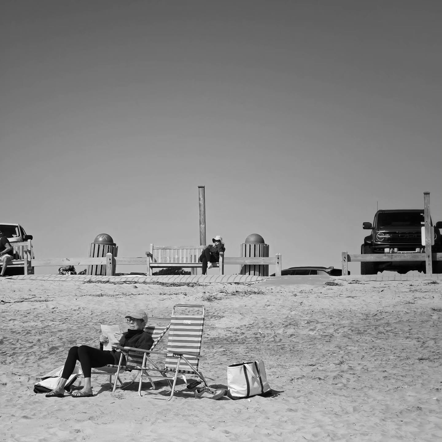 A reader settles into a striped beach chair while others take the benches along the dune fence behind.