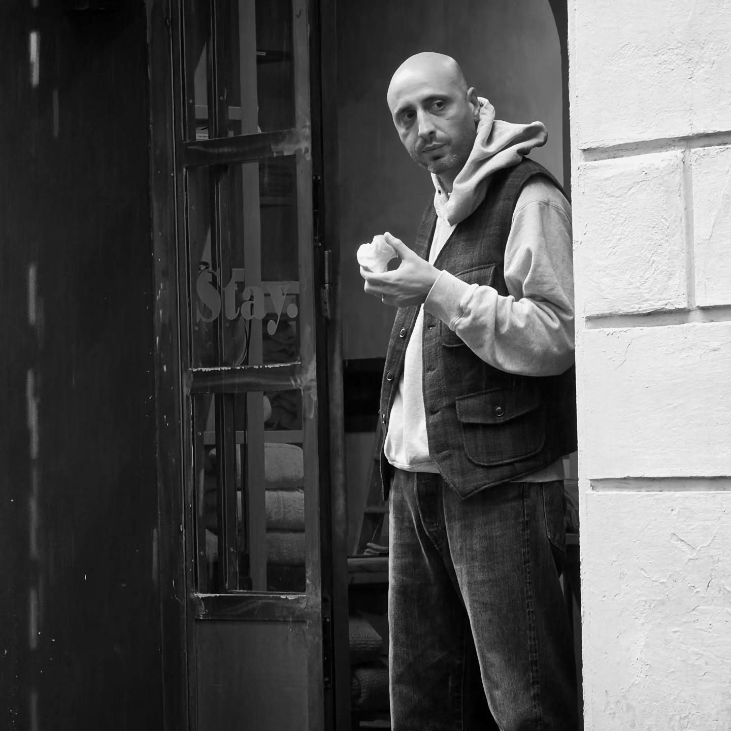 A man pauses in a shop doorway with a snack in hand, the word "Stay" etched on the glass beside him.